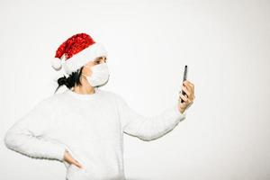 Young woman with facemask on holds cellphone high up isolated in white background. Concept of facetime and cummunication during holidays on pandemic. photo