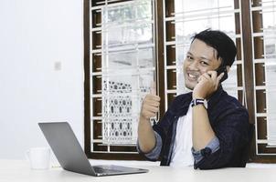 Young Asian man happy and smile what he see in the laptop when in call on smartphone photo