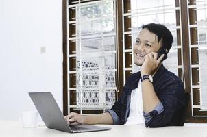 joven asiático feliz y sonríe lo que ve en la computadora portátil cuando llama al teléfono inteligente foto