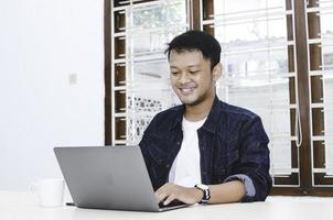 Young Asian man feeling happy and smile when work laptop on table. Indonesian man wearing blue shirt. photo