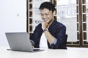 Young Asian man feeling happy and smile when work laptop on table. Indonesian man wearing blue shirt. photo