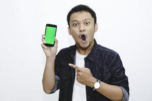 Young asian man shocked and wow with showing green screen and pointing smartphone photo