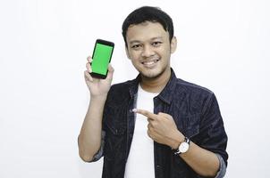 Young Asian man is standing and smiling pointing on green screen on smartphone screen on white background. photo