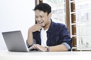 Young Asian man feeling happy and smile when work laptop on table. Indonesian man wearing blue shirt. photo