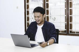 Young Asian man feeling happy and smile when work with phone and laptop on table. photo