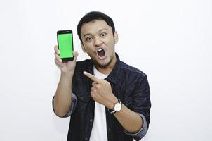 Young asian man shocked and wow with showing green screen and pointing smartphone photo