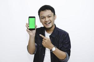 Young Asian man is standing and smiling pointing on green screen on smartphone screen on white background. photo