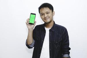 Young Asian man is standing and smiling pointing on green screen on smartphone screen on white background. photo