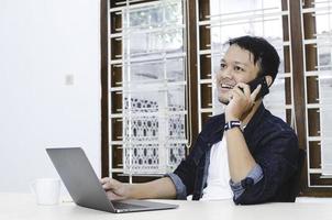 Young Asian man feeling happy and smile when work with phone and laptop on table. photo