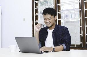 Young Asian man feeling happy and smile when work laptop on table. Indonesian man wearing blue shirt. photo