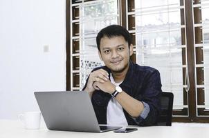 Young Asian man feeling happy and smile when work laptop on table. Indonesian man wearing blue shirt. photo