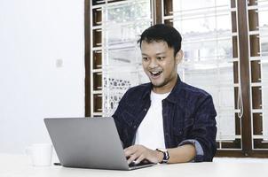 Young Asian man feeling happy and smile when work laptop on table. Indonesian man wearing blue shirt. photo