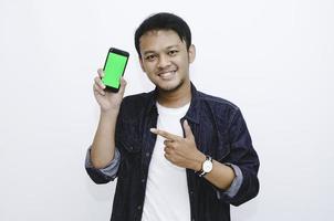 Young Asian man is standing and smiling pointing on green screen on smartphone screen on white background. photo