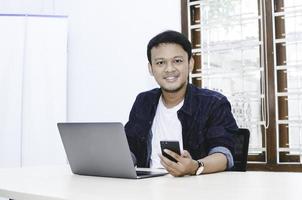 Young Asian man feeling happy and smile when work with phone and laptop on table. photo