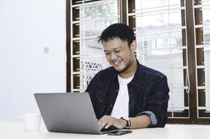 Young Asian man feeling happy and smile when work laptop on table. Indonesian man wearing blue shirt. photo