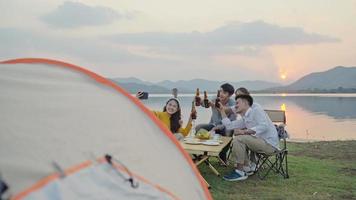 grupo de cuatro personas amigos hombres y mujeres asiáticos acampando, bebiendo cerveza, celebrando, selfie a través de un teléfono inteligente divirtiéndose y disfrutando de la tienda de tierra. área del embalse durante el tiempo de vacaciones al atardecer. video