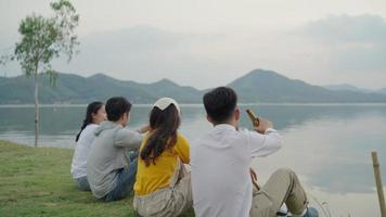 achteraanzicht. groep aziatische vier jonge vrienden mannen en vrouwen hebben een kampeerpicknick bij de rivier, ze kletsen, lachen, drinken vieren juichen bier op video