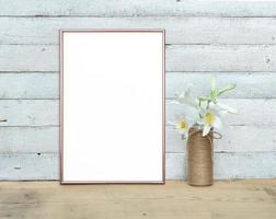 Rose Gold vertical A4 Frame mockup near a bouquet of lilies stands on a wooden table on a painted white wooden background. Rustic style, simple beauty. 3 render. photo