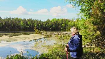 visão panorâmica mulher caucasiana loira idosa fica segurando varas nórdicas vermelhas e apreciando a natureza verde da floresta do lago