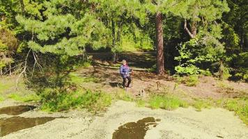 Aerial zoom out view elderly blond caucasian woman stands holding nordic walking sticks and enjoying lake and tranquil forest green nature video