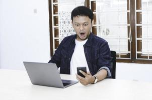 Young Asian man shocked and wow what he see in the laptop when in call on smartphone photo