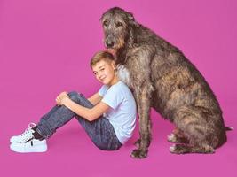 Smiling cheerful eleven years teen in white t-shirt and jeans with brown big dog on fuchsia color background in photo studio. Pets, friendship concept