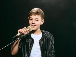 Portrait of caucasian teenager in white t-shirt, blue jeans and leather jacket with microphone singing on dark background. Hobby and glory concept photo