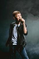 Portrait of caucasian teenager in white t-shirt, blue jeans and leather jacket with microphone singing on dark background. Hobby and glory concept photo