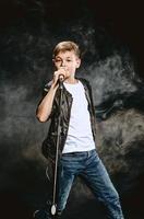 retrato de adolescente caucásico con camiseta blanca, jeans azules y chaqueta de cuero con micrófono cantando sobre fondo oscuro. concepto de hobby y gloria
