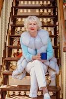 senior stylish woman in fur coat and with grey hair sitting on carousel drinking tea and enjoying life. Travel, fun, happiness, seasonal concept photo