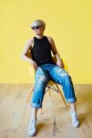 Portrait of stylish blonde young woman in black sun glasses, black shirt and jeans sitting on director chair on yellow wall background photo