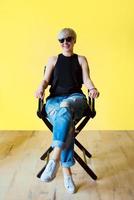 Portrait of stylish blonde young woman in black sun glasses, black shirt and jeans sitting on director chair on yellow wall background photo