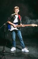 Portrait of caucasian teenager in white t-shirt, blue jeans and leather jacket with microphone and guitar singing on dark background. Hobby and glory concept photo