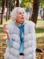 sad fashionable senior woman with grey hair in fur coat outdoor smoking cigarette. Unhealthy lifestyle, age, oldness, addiction, bad habit concept. photo