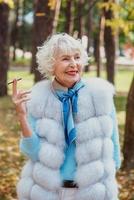 smiling senior elegant stylish fashionable woman with grey hair in fur coat outdoor smoking cigarette. Unhealthy lifestyle, age, positive vibes, oldness, addiction, bad habit concept. photo