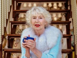 mujer elegante senior en abrigo de piel y con cabello gris sentado en carrusel bebiendo té y disfrutando de la vida. viajes, diversión, felicidad, concepto estacional. foto