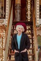 senior stylish woman in dark grey coat, hat and with grey hair standing by the carousel smiling and enjoying life. Travel, fun, pension, happiness, seasonal concept photo