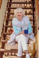 senior stylish woman in fur coat and with grey hair sitting on carousel drinking tea and enjoying life. Travel, fun, happiness, seasonal concept