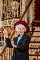 senior stylish woman in dark gray coat, hat and with grey hair standing by the carousel smiling and enjoying life. Travel, fun, pension, happiness, seasonal concept photo