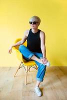 Portrait of stylish blonde young woman in black sun glasses, black shirt and jeans sitting on director chair on yellow wall background photo