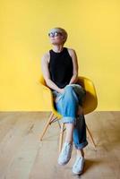 Portrait of stylish blonde young woman in black sun glasses, black shirt and jeans sitting on director chair on yellow wall background photo