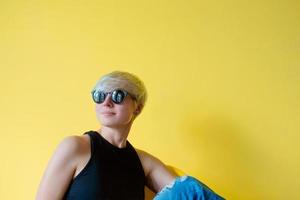 retrato de una joven rubia elegante con gafas de sol negras, camisa negra y jeans sentada en una silla de director con fondo de pared amarilla foto