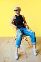 Portrait of stylish blonde young woman in black sun glasses, black shirt and jeans sitting on director chair on yellow wall background photo