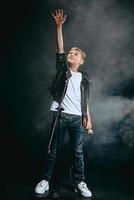 Portrait of caucasian teenager in white t-shirt, blue jeans and leather jacket with microphone singing on dark background. Hobby and glory concept photo