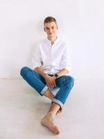 handsome guy in white shirt and blue jeans sitting on a floor indoor. photo