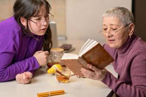 abuela y nieta beben té sentadas en la mesa y leen un libro. foto