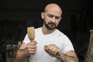 white bald carpenter with beard in white t-shirt works as a chisel and a mallet photo