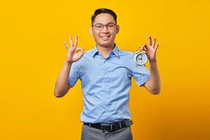 Portrait of cheerful handsome Asian young man in glasses showing alarm clock and showing okay gesture with finger isolated on yellow background. Time management lifestyle concept photo