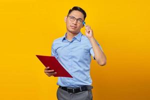Portrait of young handsome Asian man scratching head with pen, thinking of ideas and holding document folder isolated on yellow background photo