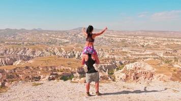 statico rallentatore vista fidanzata sedersi sulle spalle del ragazzo godersi il viaggio in Cappadocia scenica e mostrare il segno di amore sul bordo della scogliera. una coppia innamorata si trova sul bordo di una scogliera in Turchia video